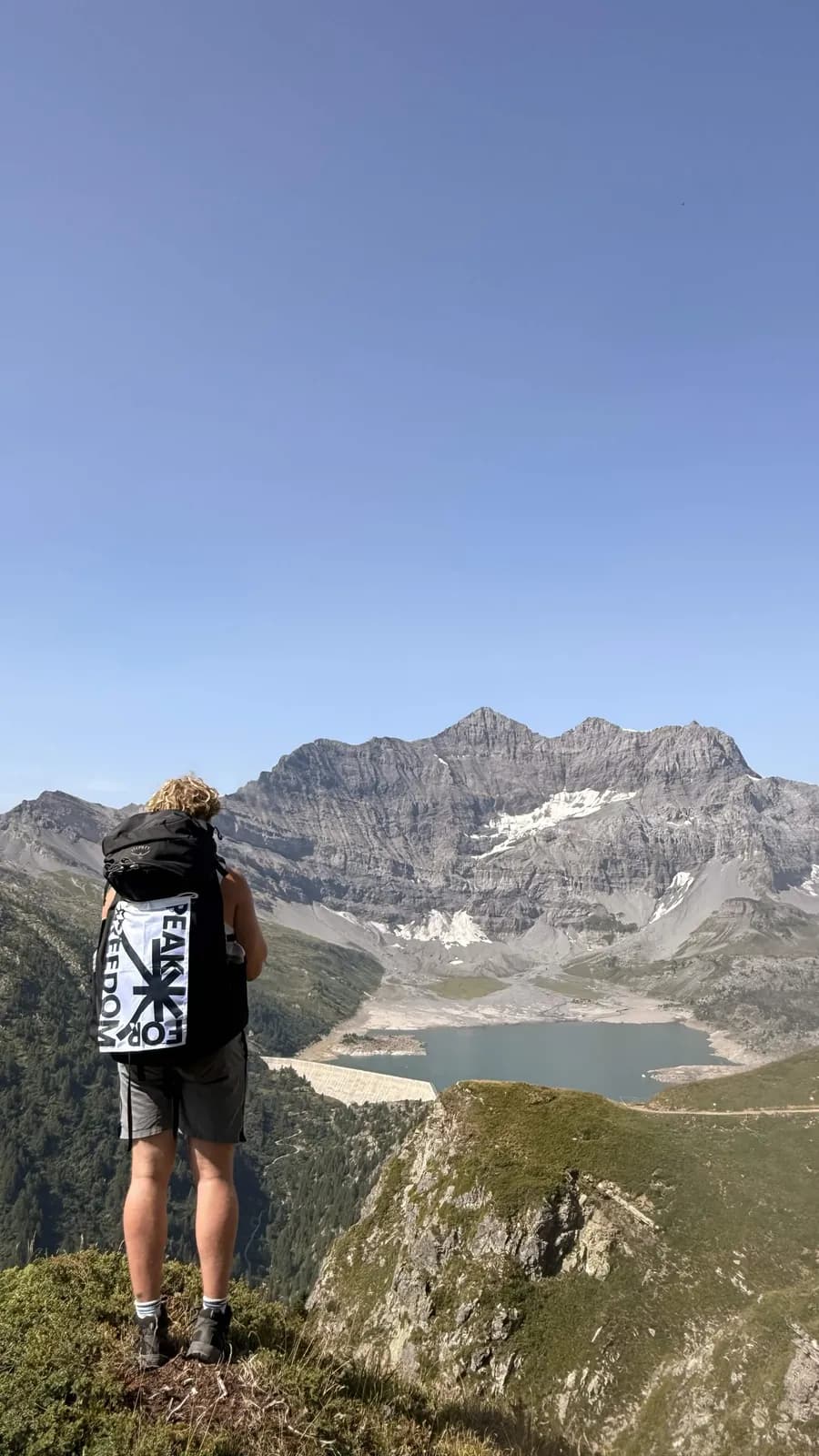 Hiker seen from behind with the Peak For Freedom flag on a backpack, looking over a turquoise mountain lake and a snow-streaked peak