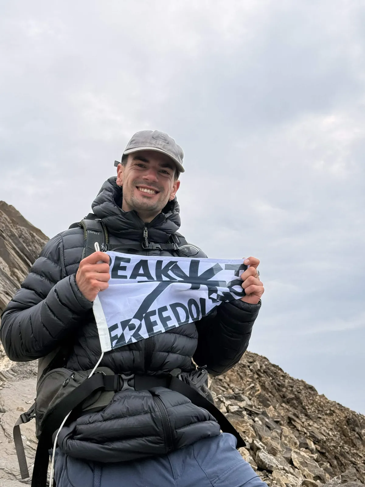 Niek on a rocky ridge in a down jacket and cap, smiling and holding the Peak For Freedom flag