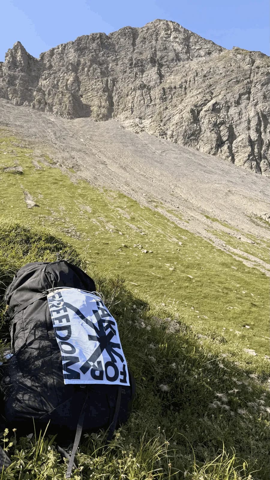 Backpack with the Peak For Freedom flag lying on a grassy slope below a grey scree face