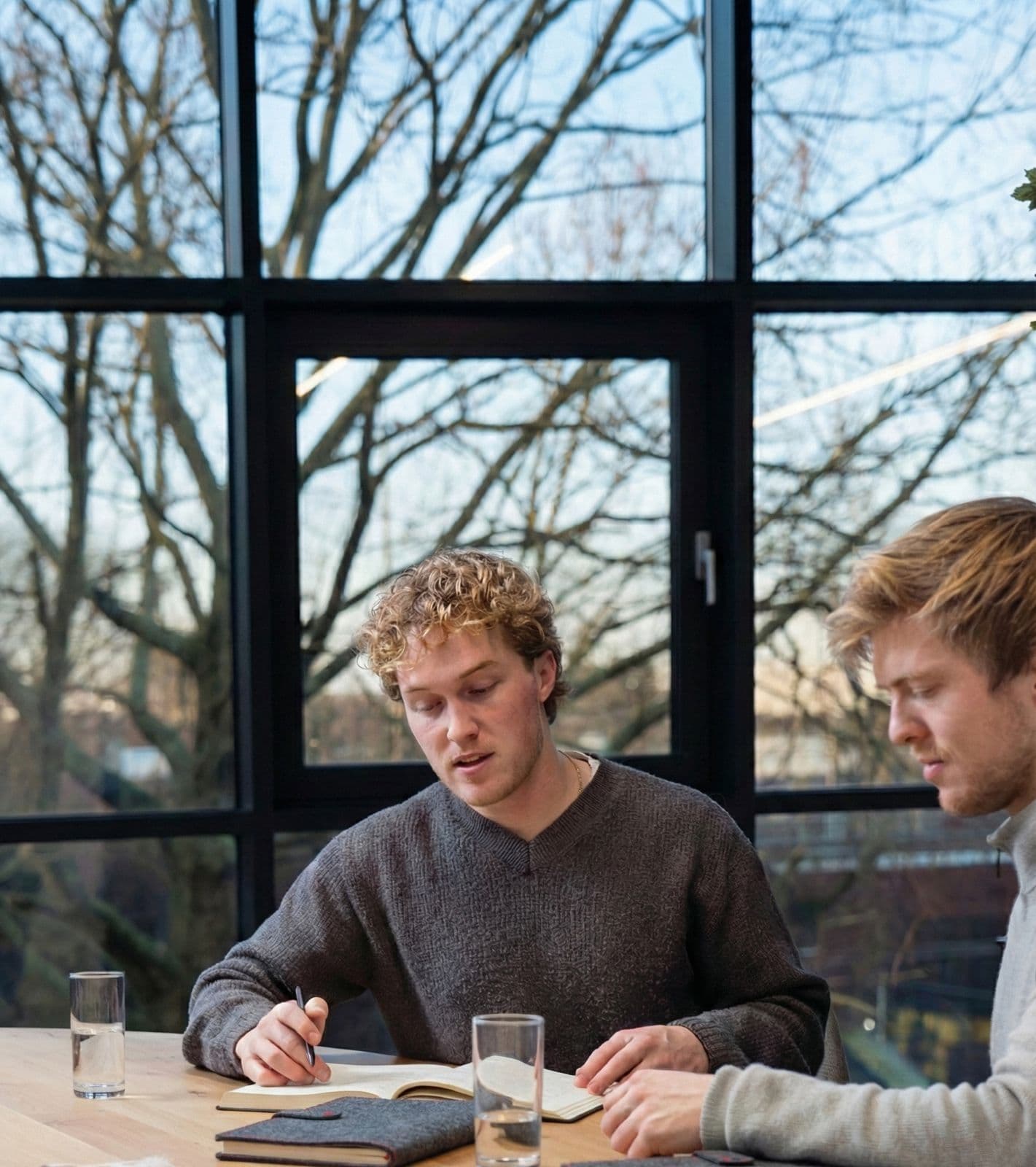 Twee mannen aan een houten tafel voor een groot raam, een van hen schrijft in een notitieboek