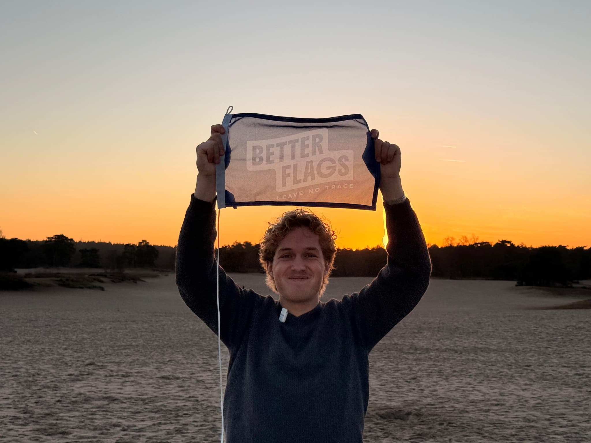 Man holding a Better Flags flag above his head on a sandy plain at sunset